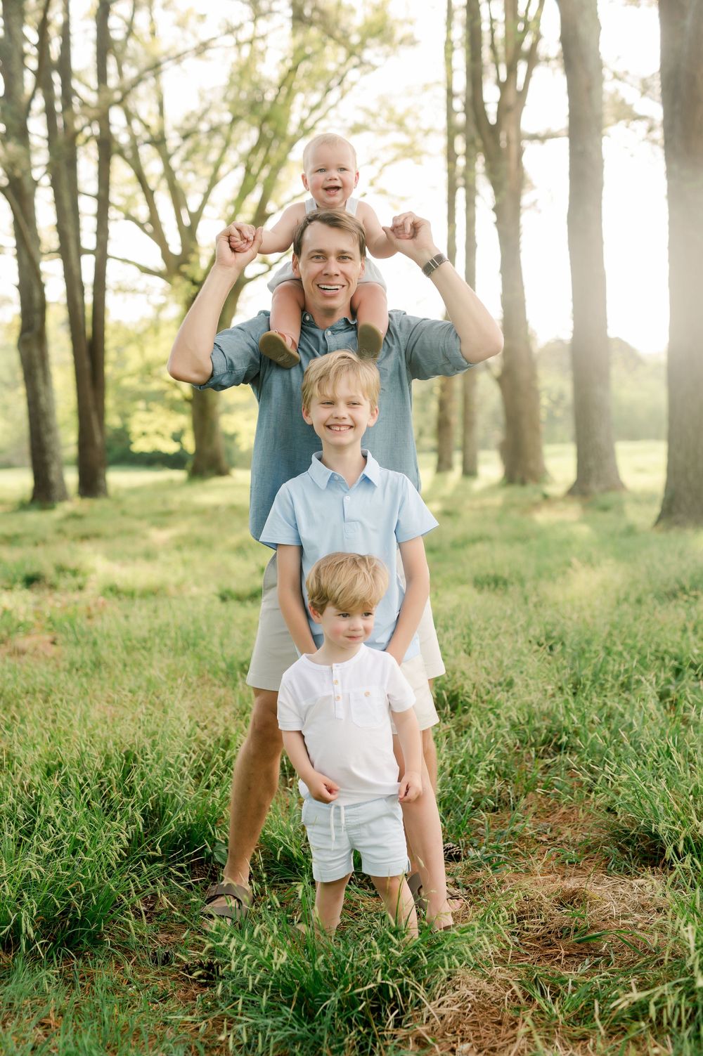 A parent carries a toddler on shoulders while standing outdoors with two other children in a sunlit wooded area.