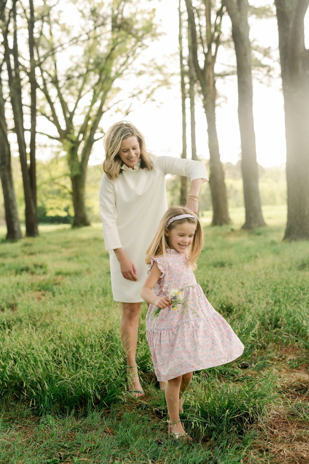 Two people share a tender moment dancing together in a sunlit forest setting wearing white and pink dresses.