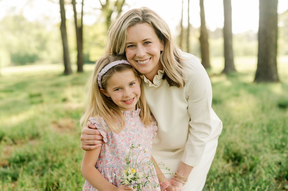 A parent and child smile together while holding wildflowers in a sunlit forest setting during spring.