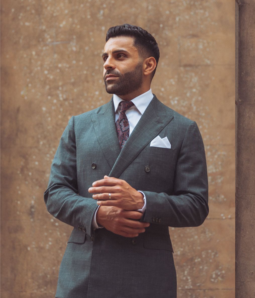 Groom in a dark grey double-breasted suit and patterned tie poses against textured brown wall.