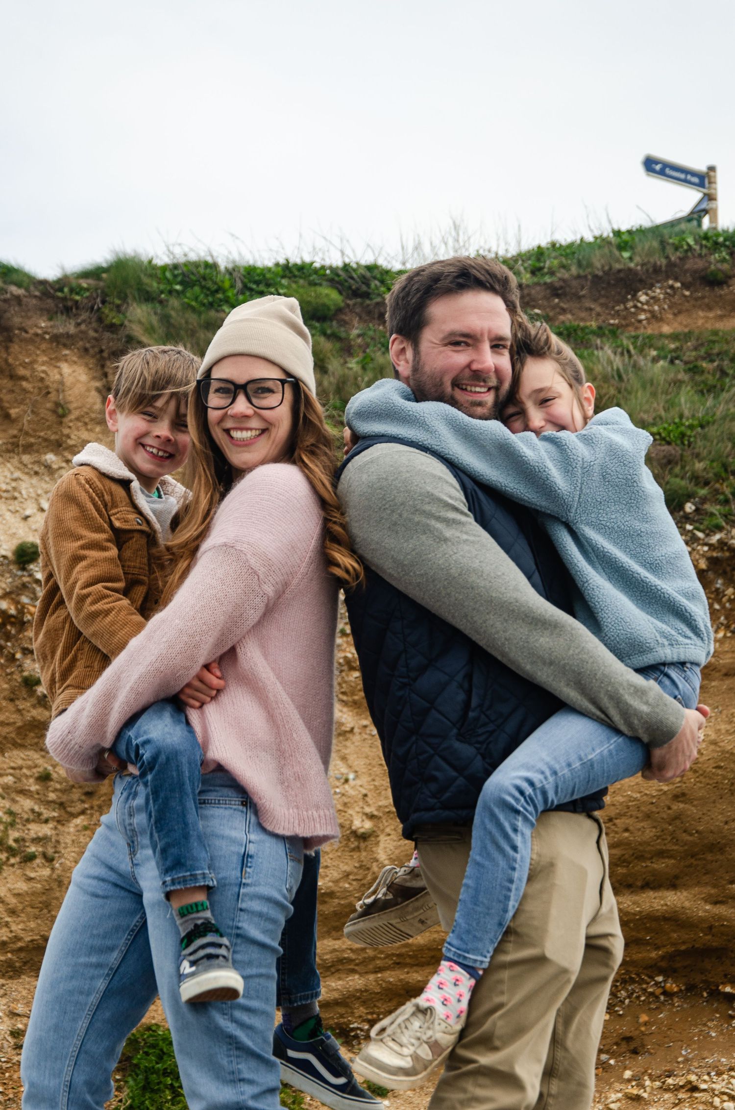 Family members carrying each other piggyback style against an outdoor hillside backdrop.