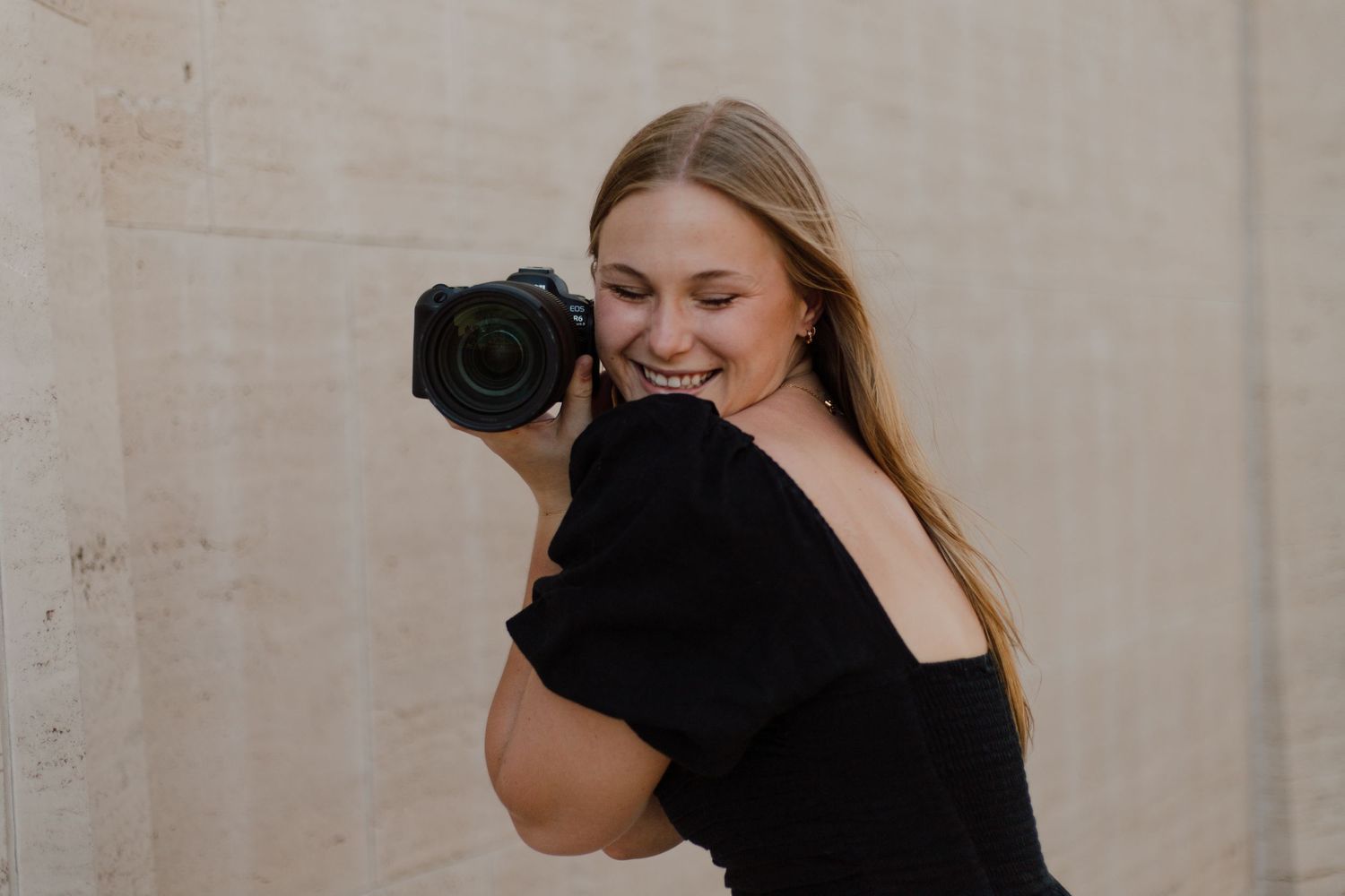 A photographer wearing a black top smiles while holding a professional DSLR camera against a light colored wall.