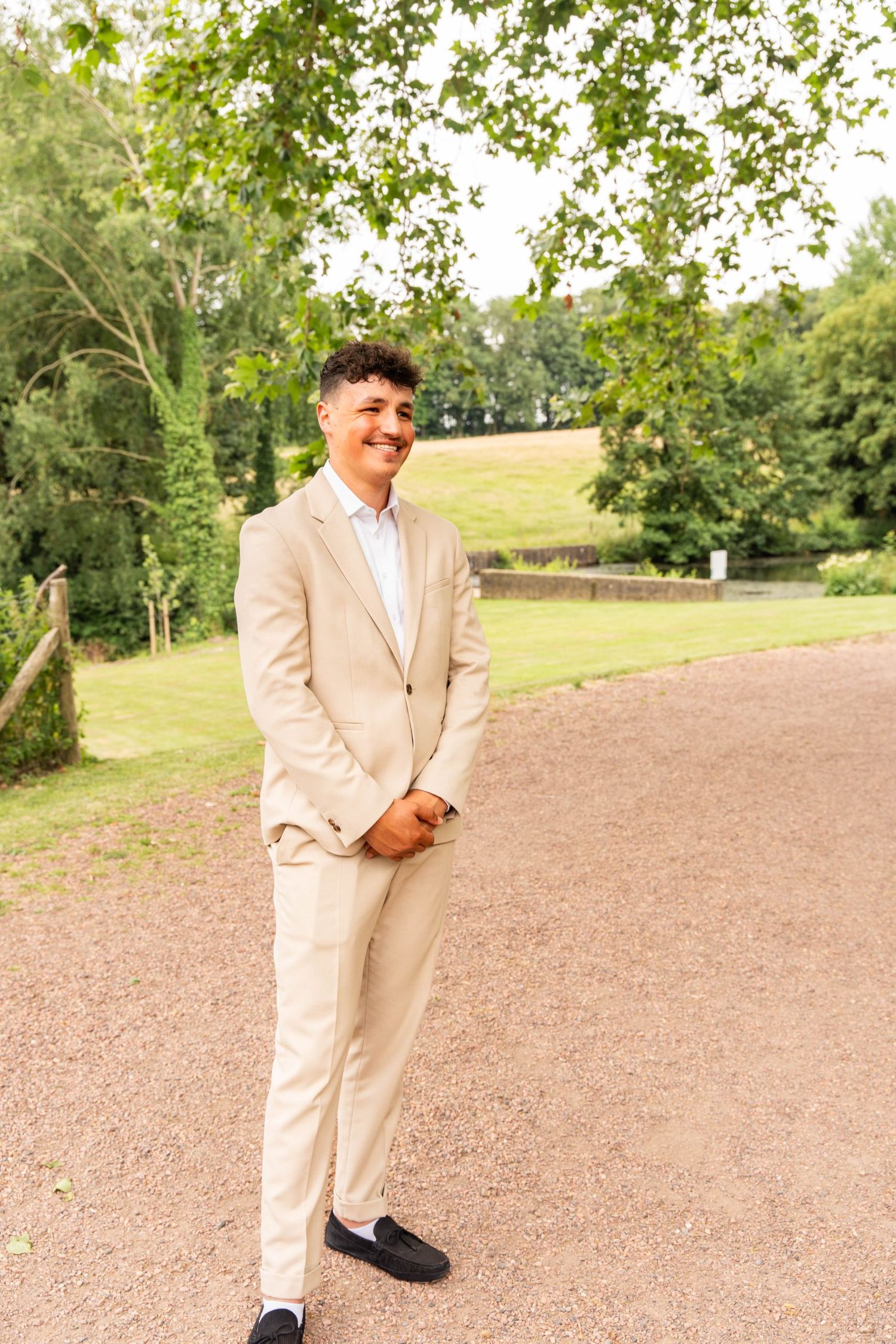A person in a beige suit poses on a dirt path surrounded by green trees.