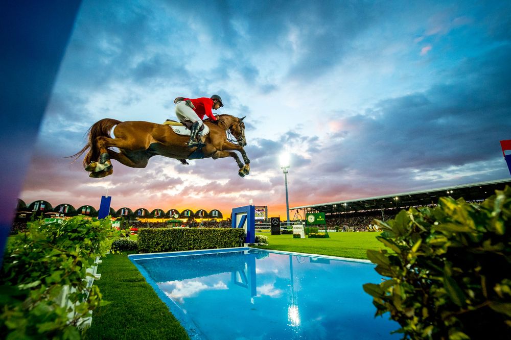 Horse and rider jumping over water obstacle at sunset with swimming pool and stadium in background.