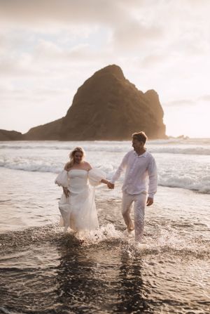 Newlyweds play in the ocean waves at sunset with a rocky coastline in the background.