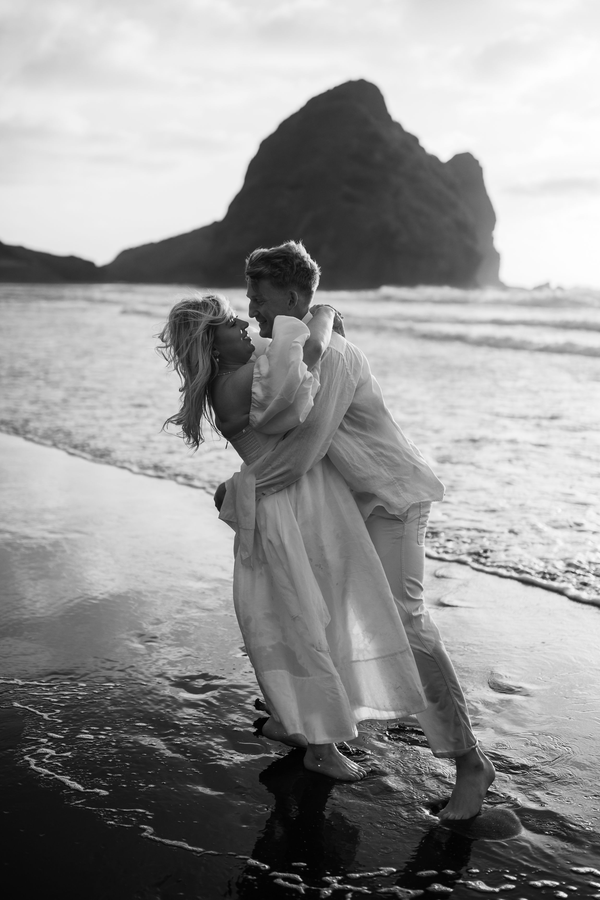 Black and white photo of couple playing in ocean waves with dramatic coastal backdrop.