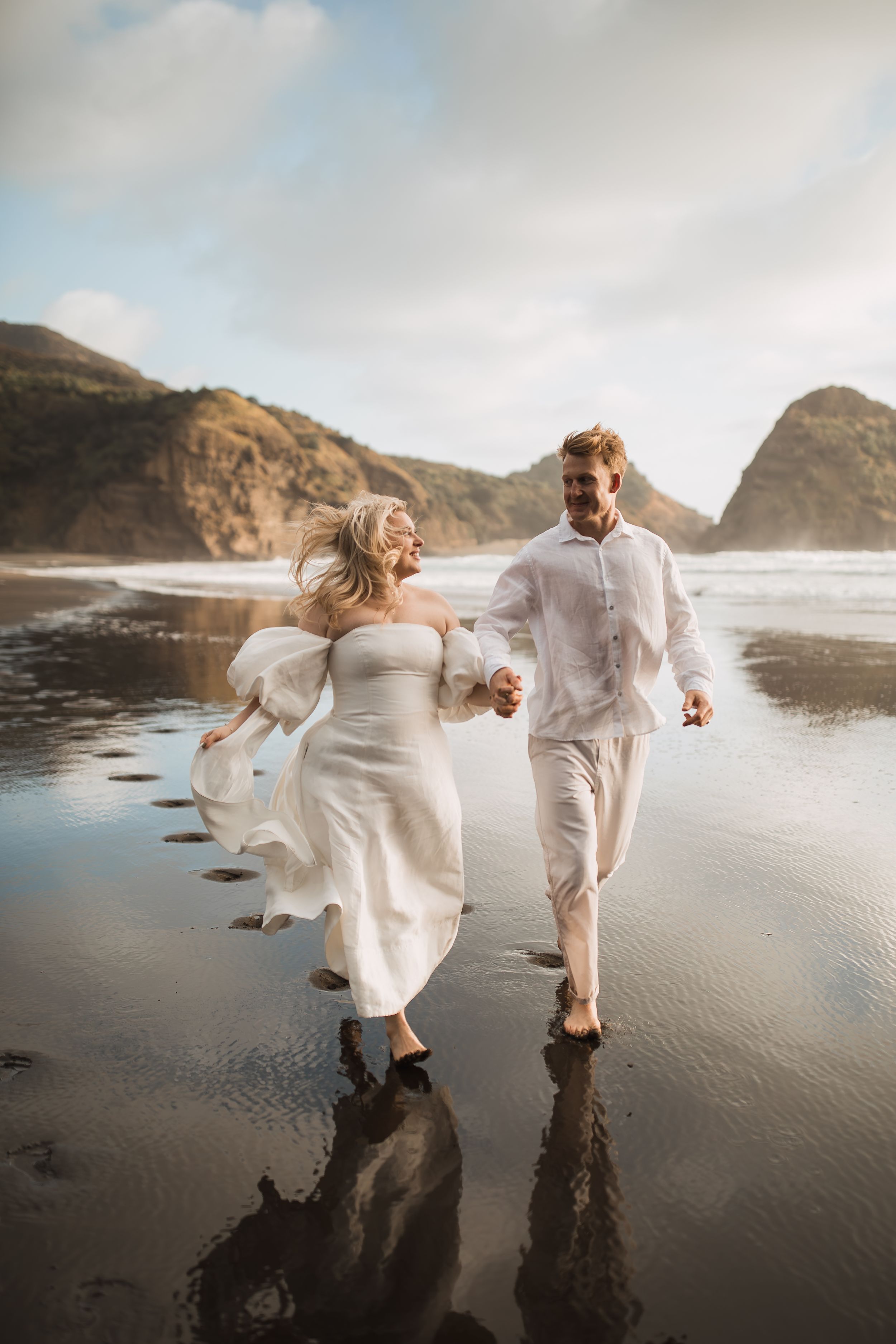 A couple in white outfits runs playfully along a dramatic black sand beach at sunset.