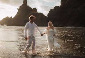Couple dressed in white walks hand in hand on a rocky beach with cliffs in the background.