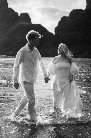 Black and white photo of two people splashing through shallow ocean water in white clothing.