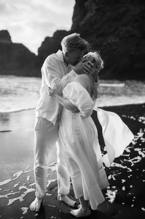 Black and white image of a couple in white attire embracing on a rocky shoreline.