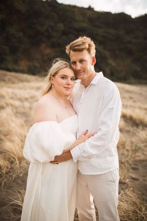 A romantic couple in white outfits embraces in golden sunlit coastal grassland.