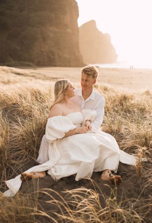 A couple in white clothing sits intimately together in coastal grass during golden hour.
