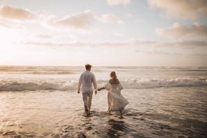 Two people in white clothing walk along a misty beach at sunset.