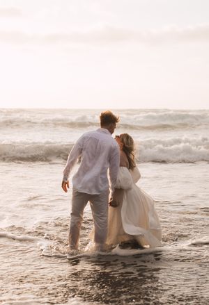 Couple in white clothing sits together on a beach at sunset as waves crash around them.