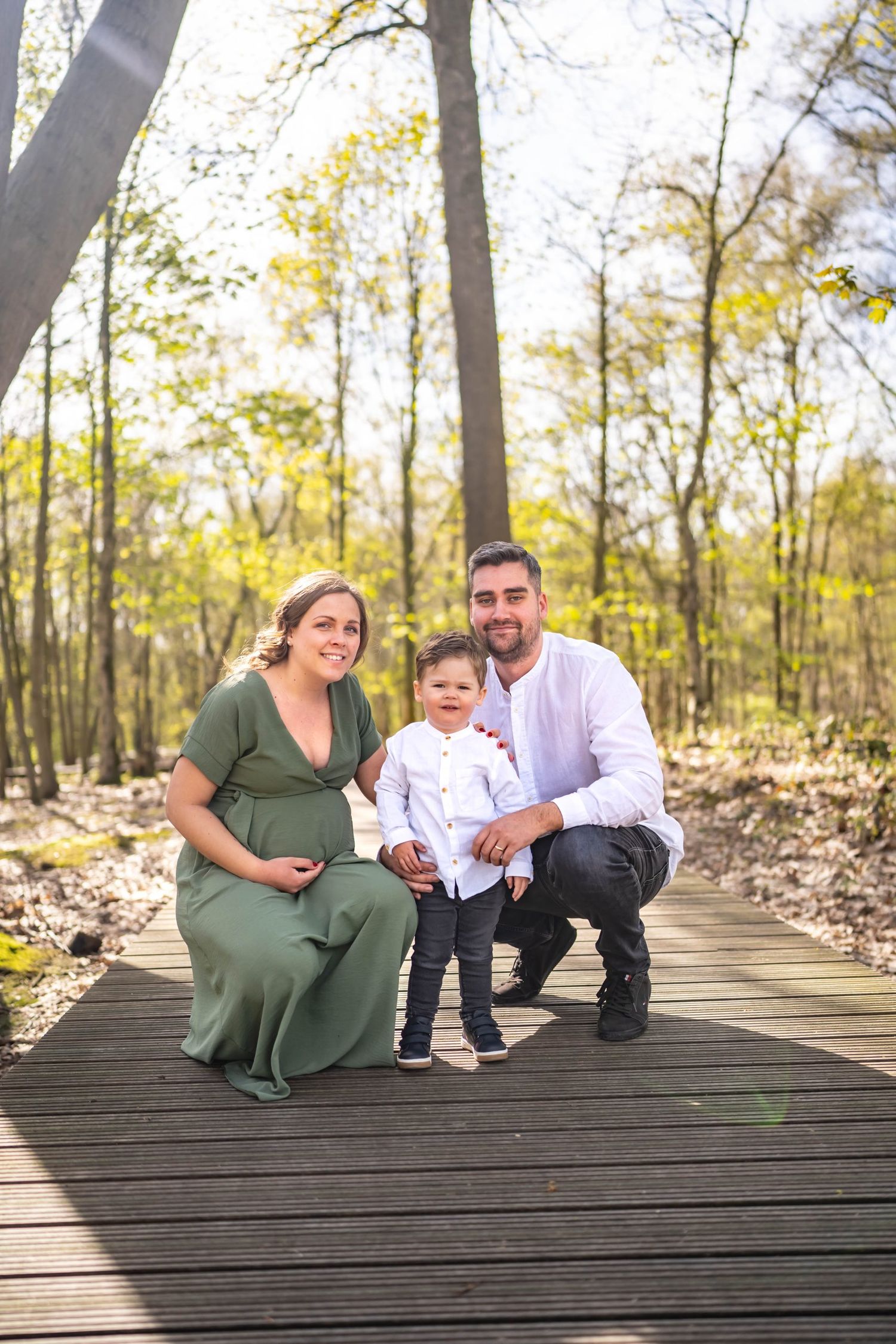 A family poses together on a wooden boardwalk in a sunlit forest setting.