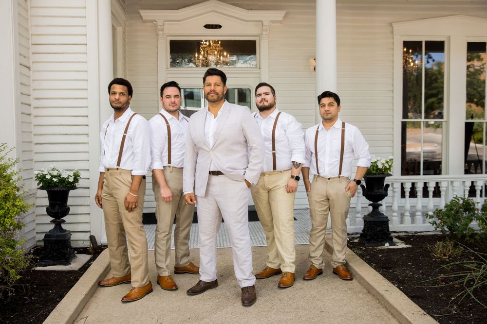 Wedding party in coordinated attire of khaki pants, suspenders, and white shirts pose on white porch steps.