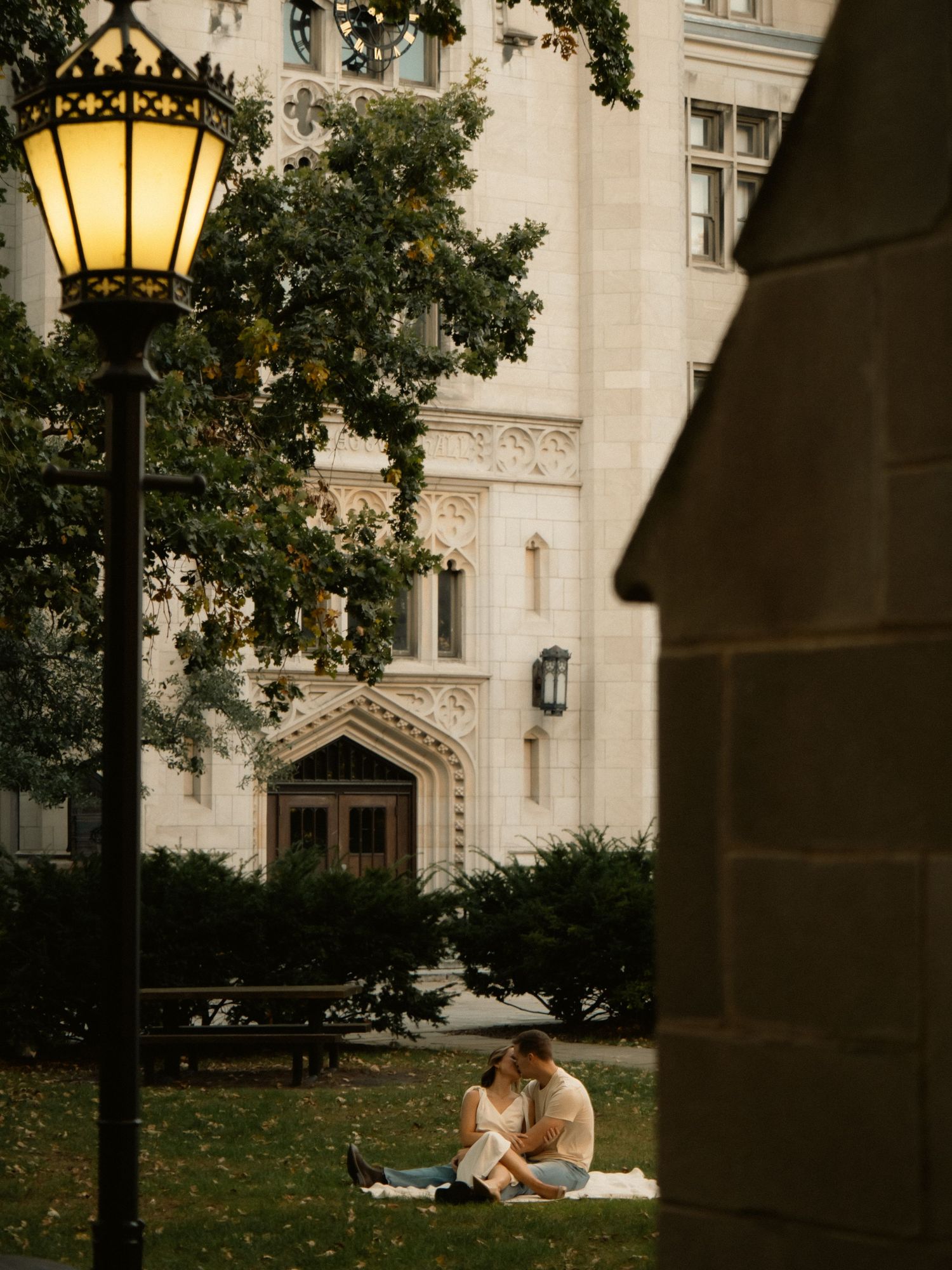 A couple shares an intimate moment on a blanket near a lit lamppost by a gothic building.