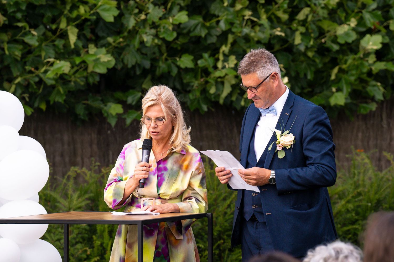 Outdoor ceremony participants reading from papers during formal gathering in garden setting.