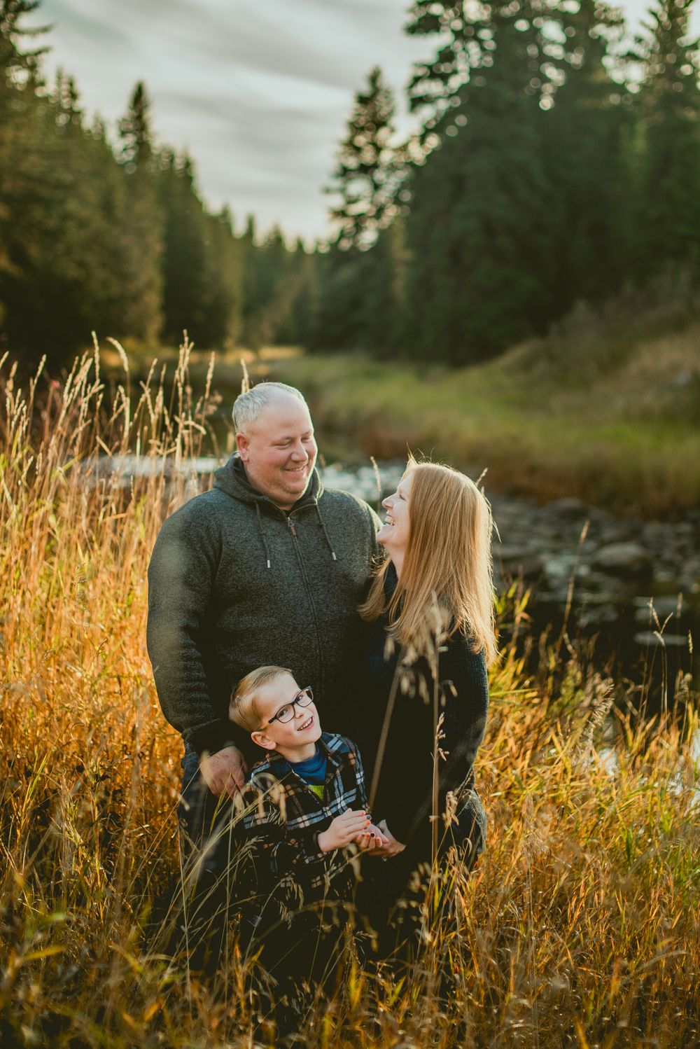 Family enjoying a golden autumn evening together in tall grass by a stream with evergreen forest in background. Gibbons Family Photography