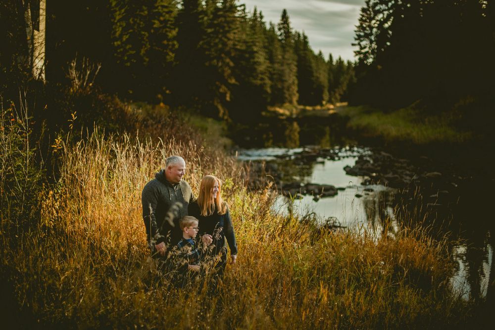 Silhouettes sit together in golden grass beside a peaceful river at sunset with evergreen forest in background.