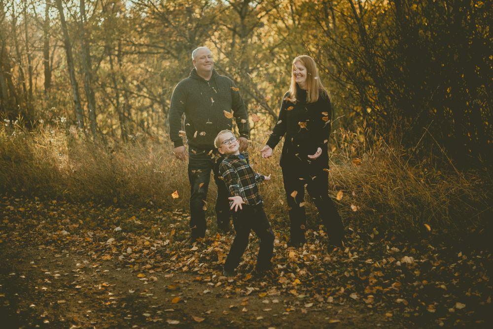 A family walks together on a golden autumn path through the woods during sunset, surrounded by fallen leaves. Gibbons Family Photography