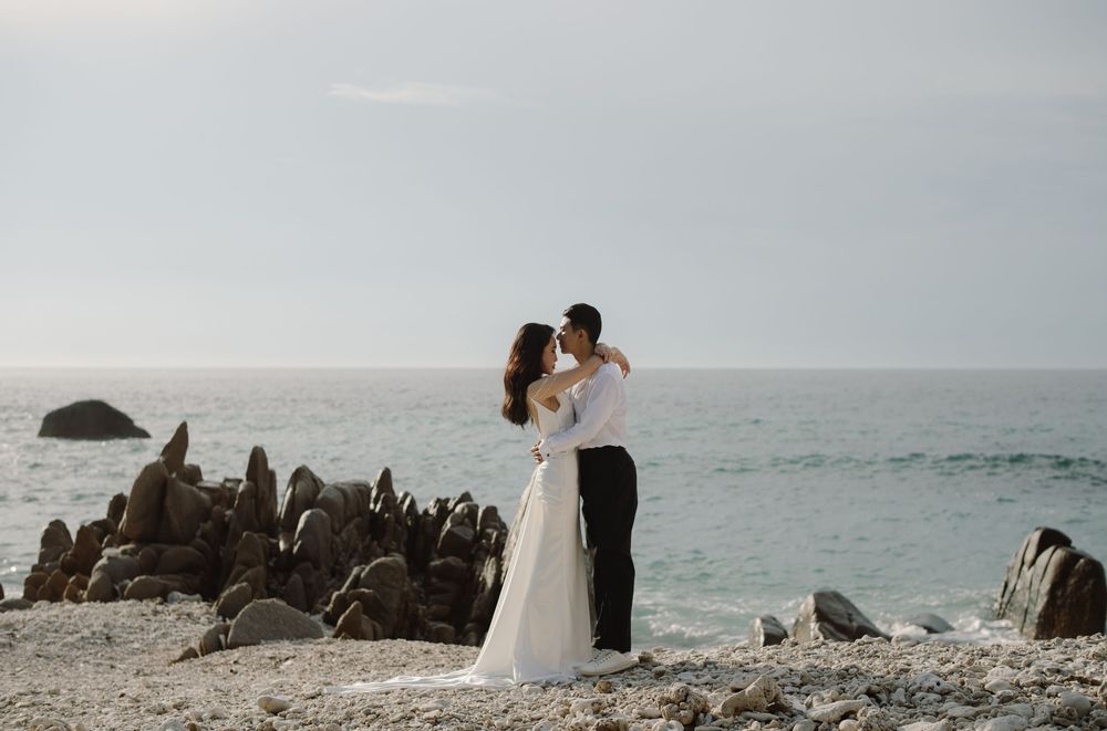 A romantic couple embraces on a rocky beach overlooking the ocean at sunset.