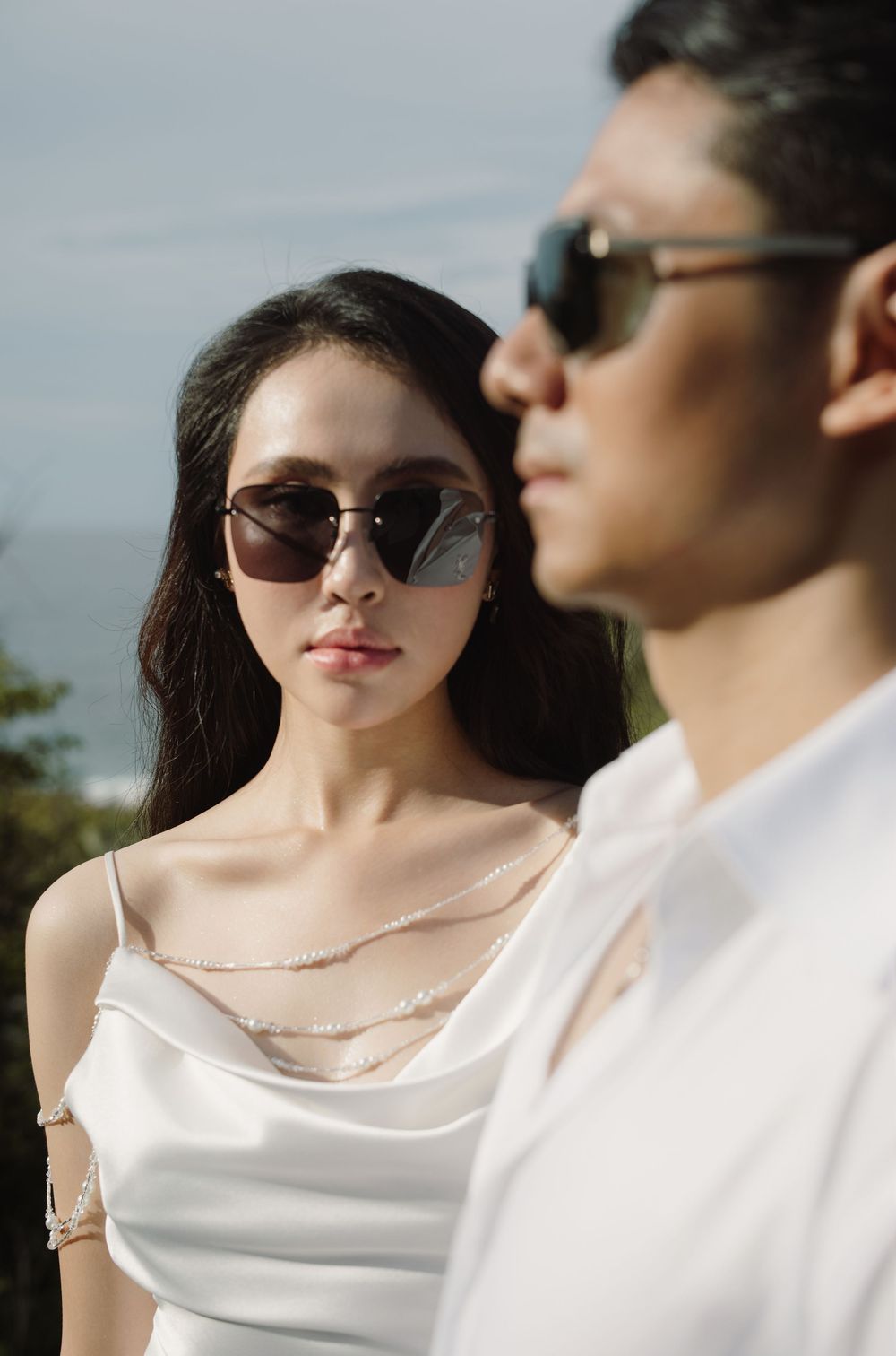 Contemporary wedding couple in white attire and sunglasses posing outdoors against overcast sky.