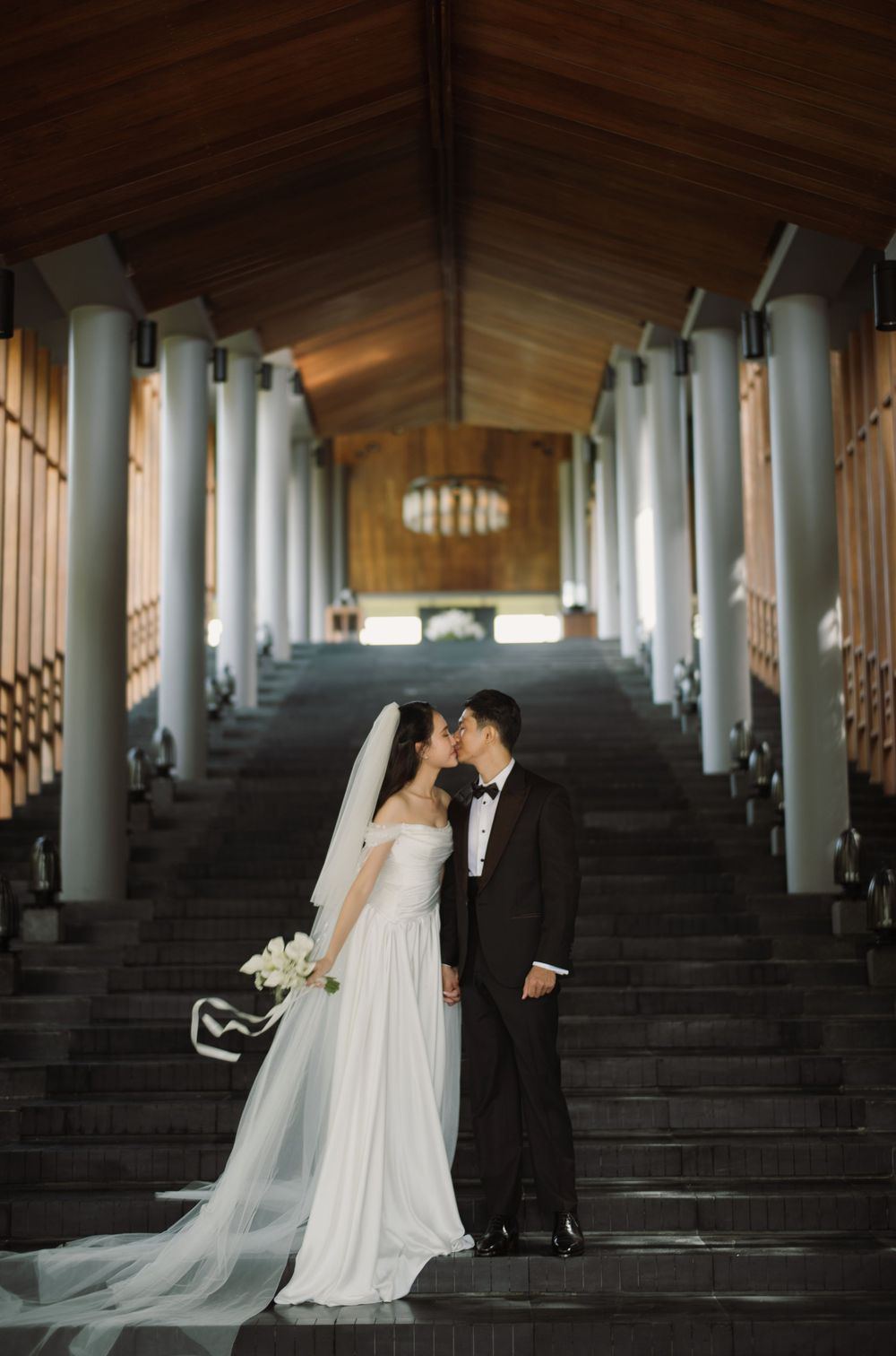 Romantic wedding photo of a couple on grand stone steps flanked by white pillars in an elegant venue.