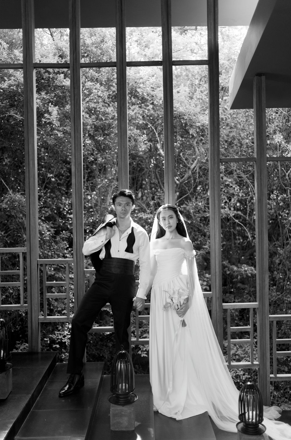 A couple in formal wedding attire poses in a sunlit room with large windows and greenery visible outside.