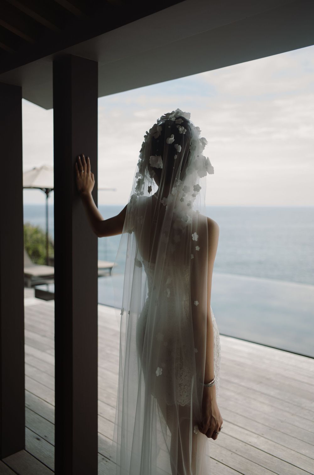 A bride in a flowing white dress and veil stands against columns overlooking an ocean view deck.