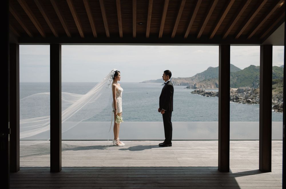 Two silhouettes stand facing each other on a seaside deck with mountains in the background.