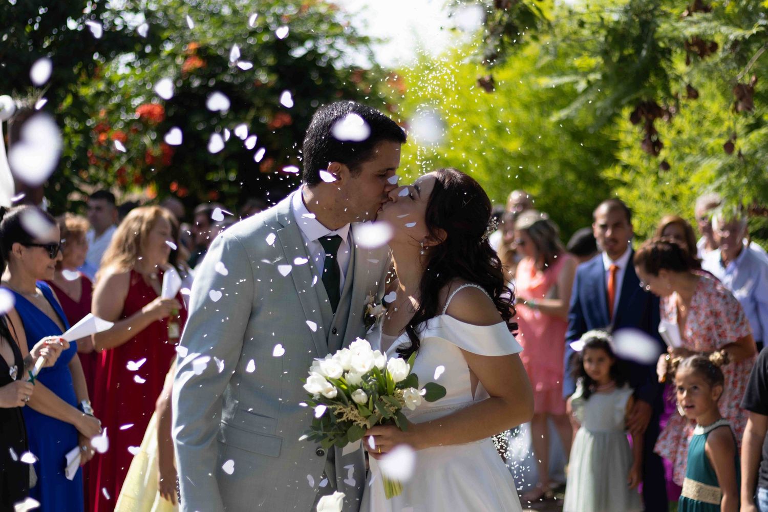 Wedding couple sharing kiss under shower of white confetti with guests celebrating.