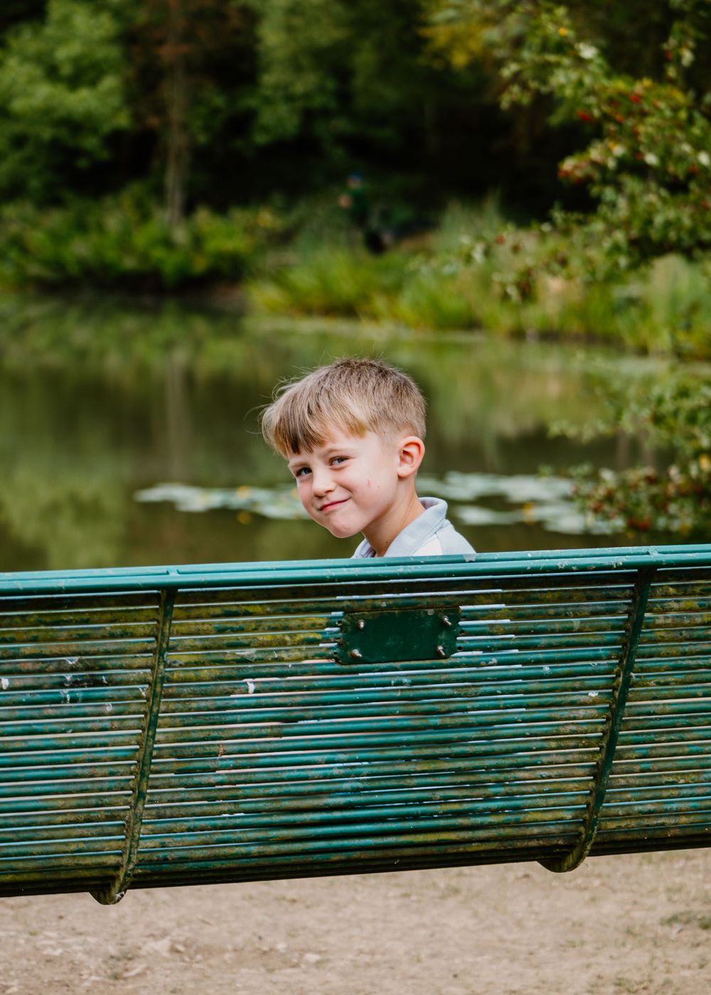 Family Photoshoot - A child peeks playfully over a turquoise park bench near a pond with lily pads in the background.