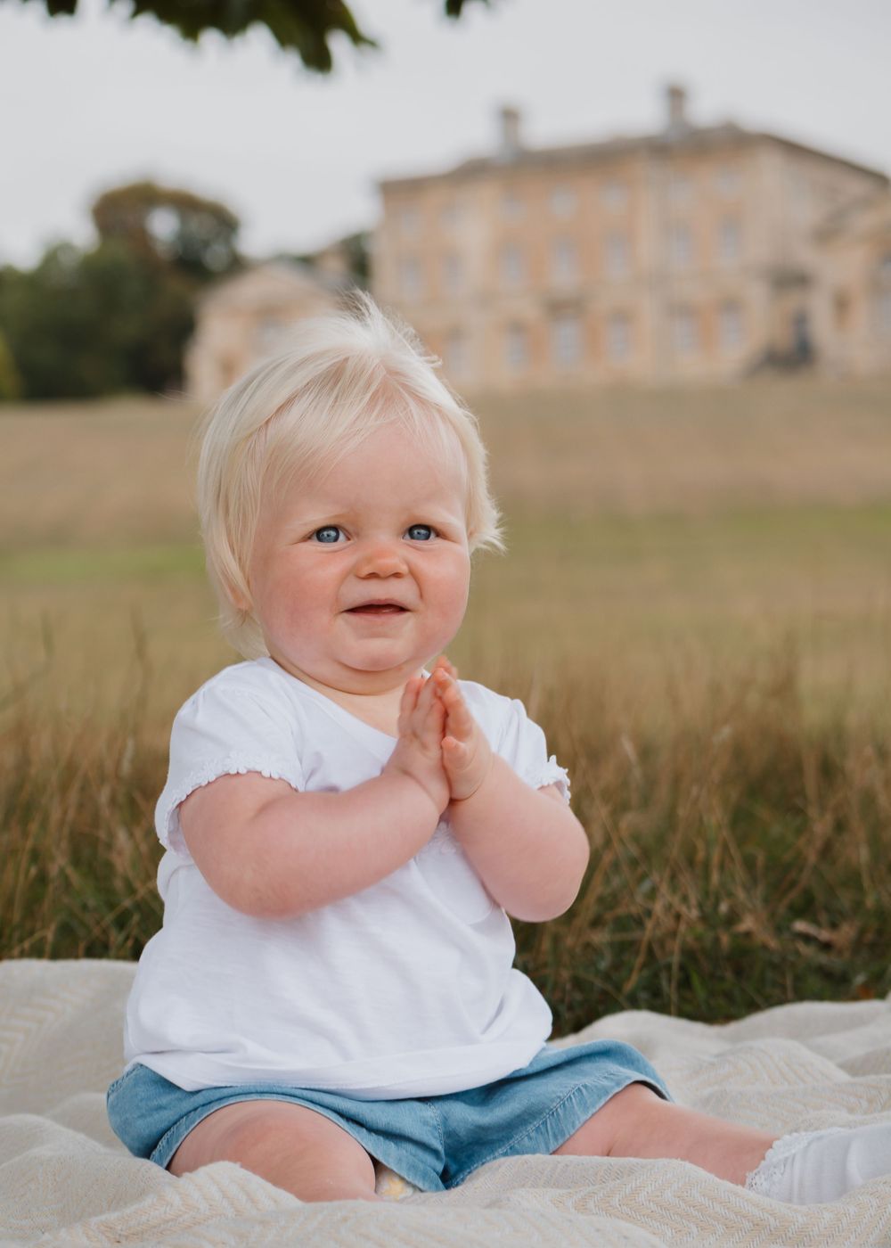 Family Photoshoot - Cusworth Hall - Baby in white top and blue shorts sits on blanket outdoors with historic building in background.