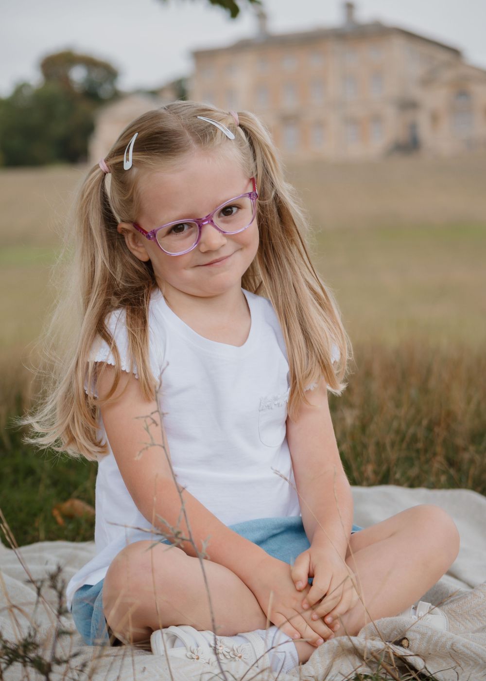 Family Photoshoot - Cusworth Hall - Young child in glasses and pigtails sits on blanket in grassy field.