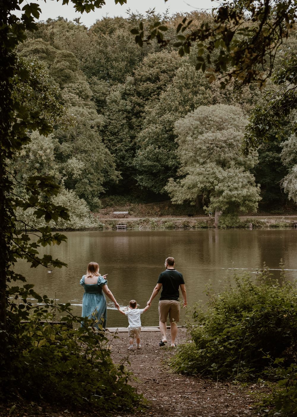 Family Photoshoot - Newmillerdam - Family silhouettes walking along a peaceful lake shore surrounded by trees.