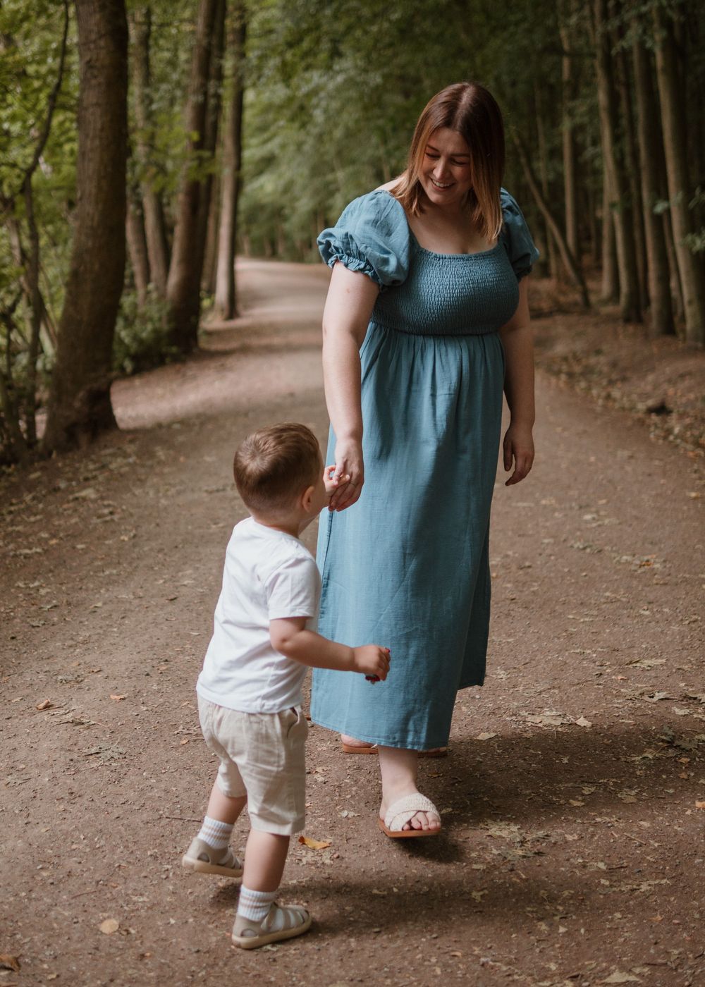 Family Photoshoot - Newmillerdam - A parent in a blue dress holds hands with a toddler while walking along a peaceful forest path.