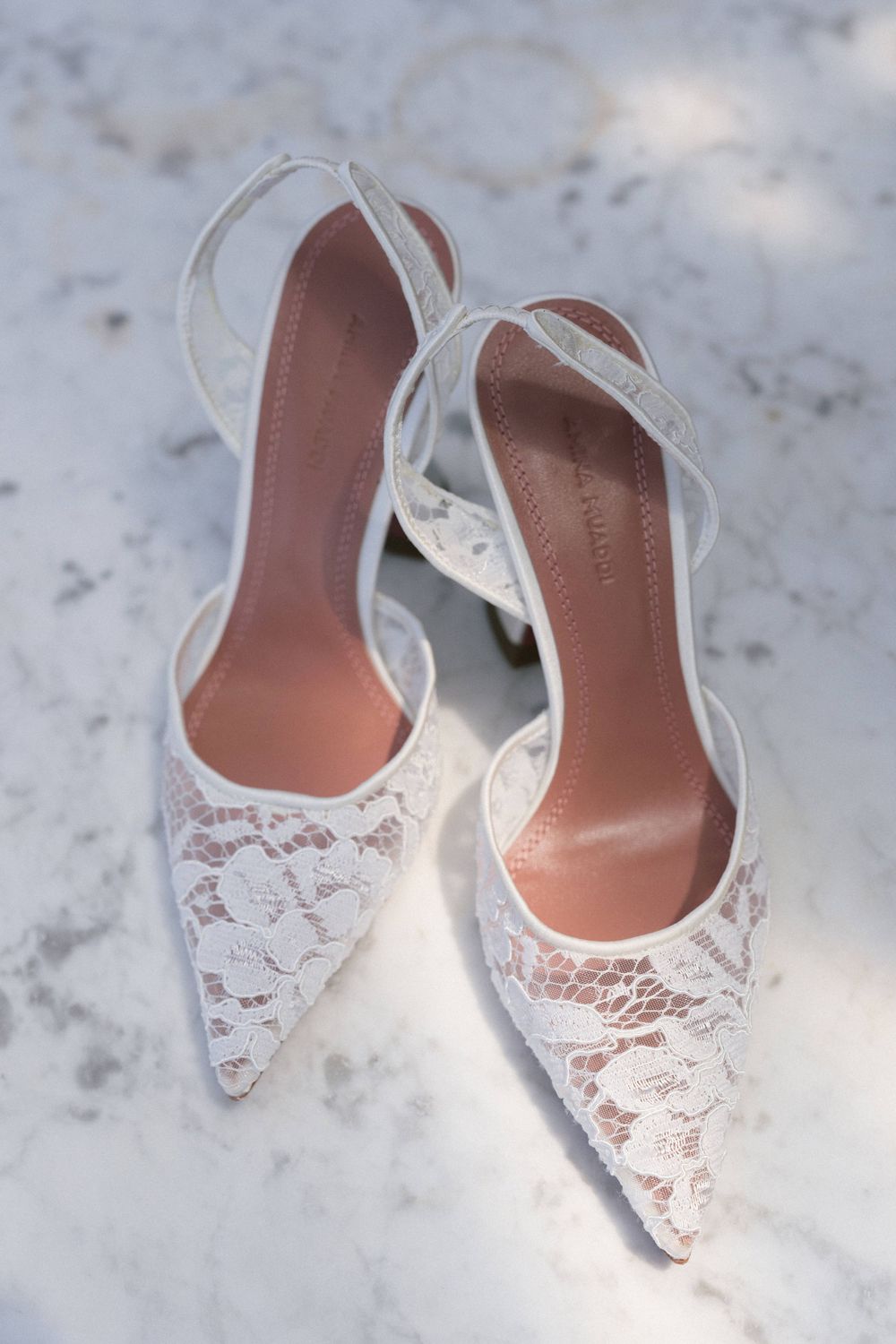 Elegant white lace wedding heels with pointed toes displayed on a marble surface.