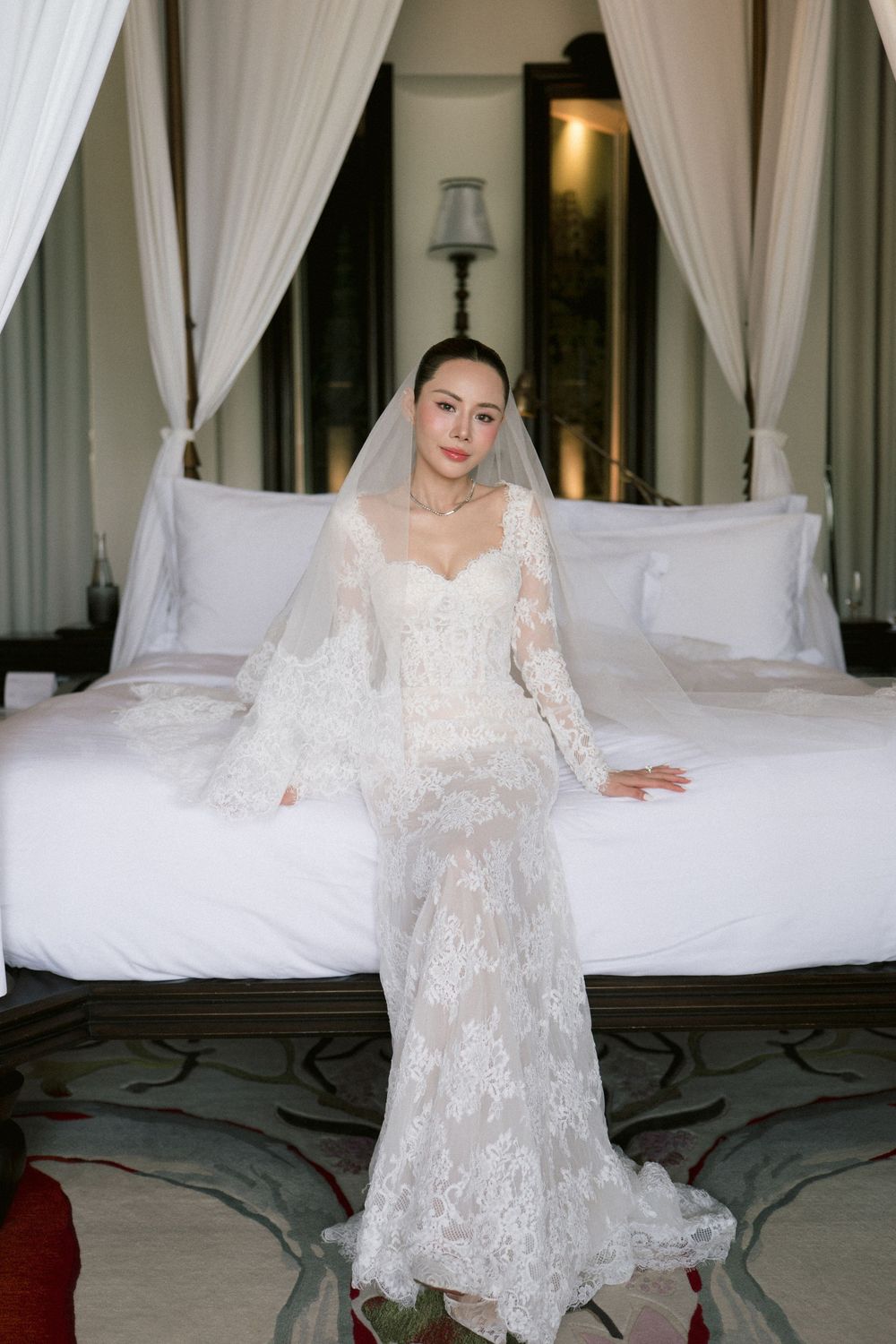 A person in an elegant long-sleeved white lace wedding dress poses gracefully in a luxurious hotel room.