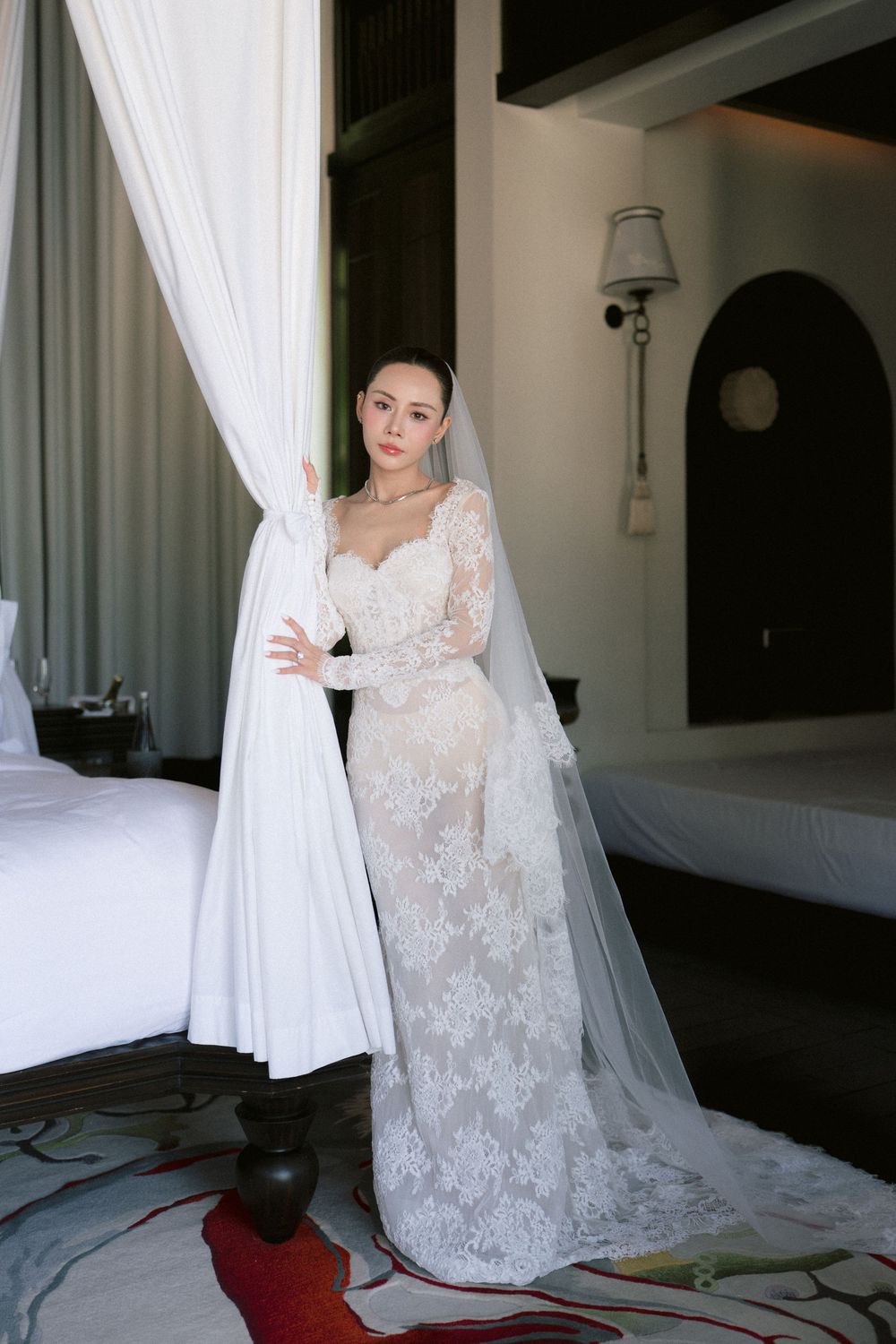 A bride in an elegant white lace wedding gown stands in a bright hotel room with flowing curtains.