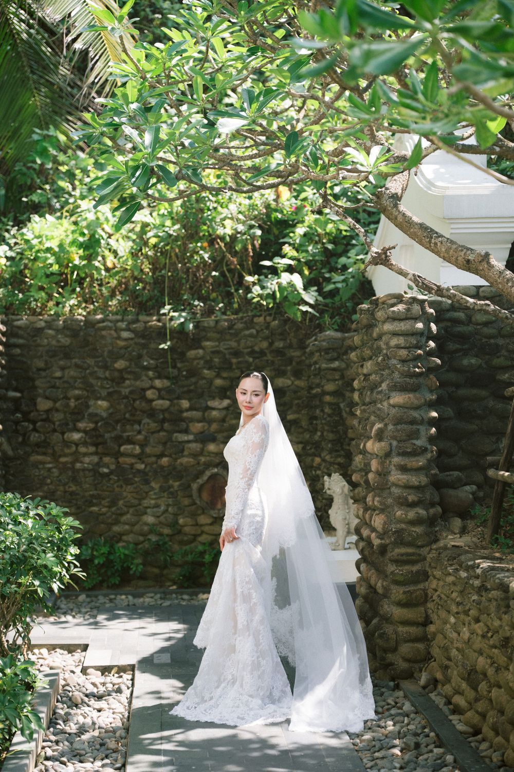 A white wedding dress with lace details photographed in a garden setting with stone walls.