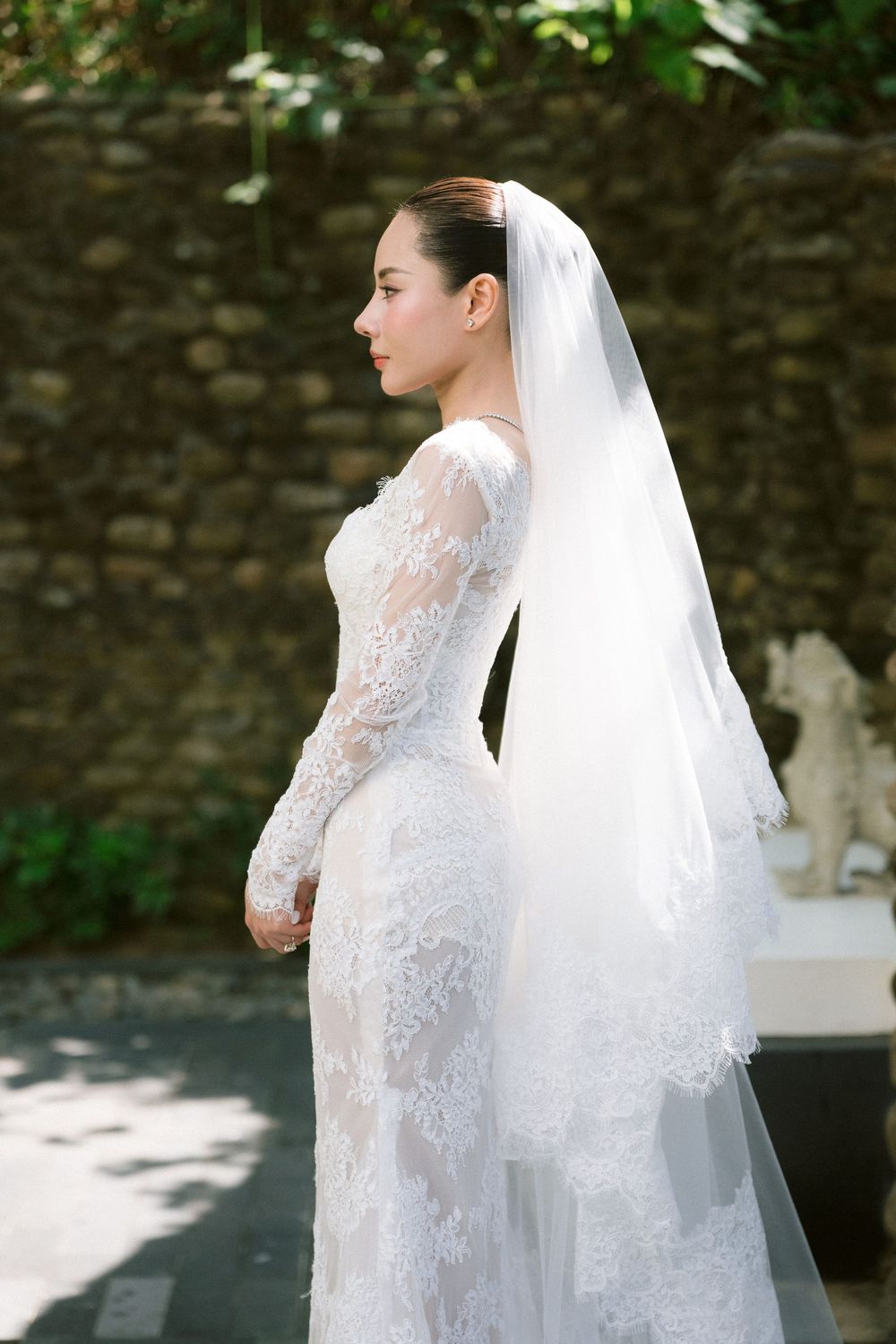 A bride in a long-sleeved lace wedding dress and veil poses against a stone wall in an outdoor garden setting.