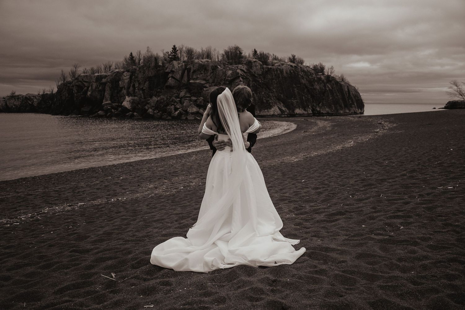 A bride in a flowing white wedding dress stands alone on a beach with a forested island in the moody background.