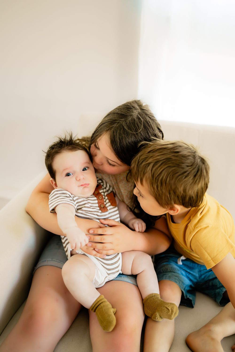 Siblings sharing tender moments with a baby on a white couch.