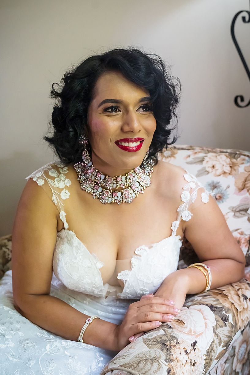 Bride wearing white lace dress with red lipstick lounges on floral upholstered furniture in elegant boudoir portrait series.