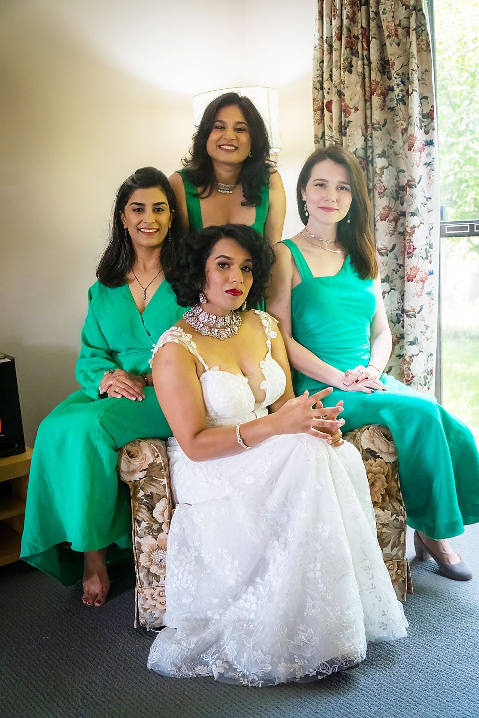 Four women in coordinating emerald green dresses and a white wedding gown pose together on floral furniture.