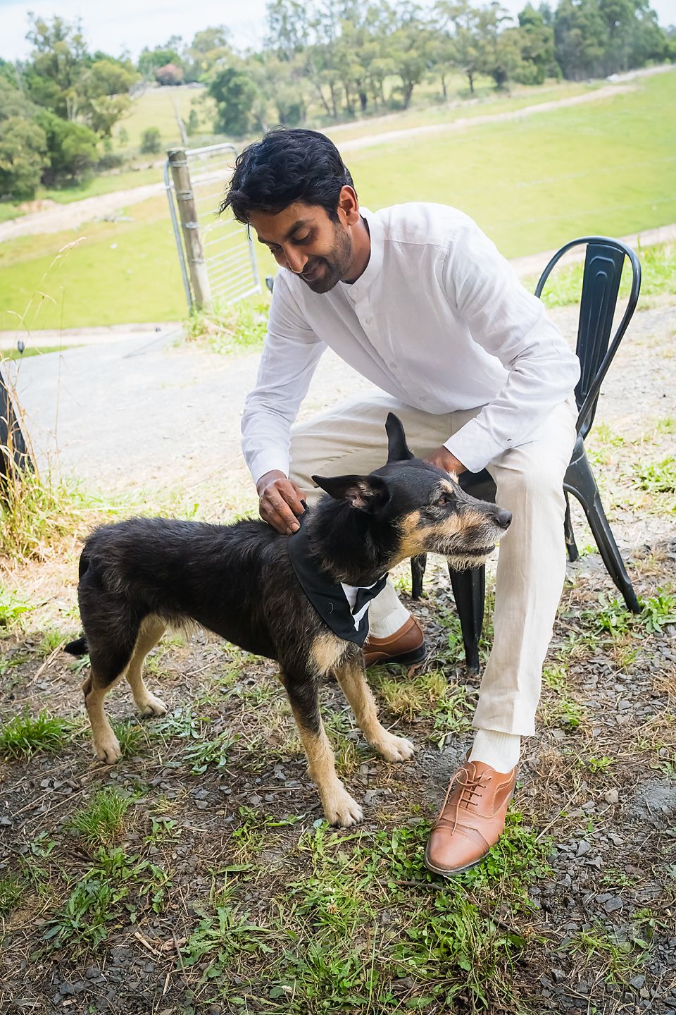 A person in white attire pets a black and tan dog while sitting on an outdoor chair with rolling hills in background.