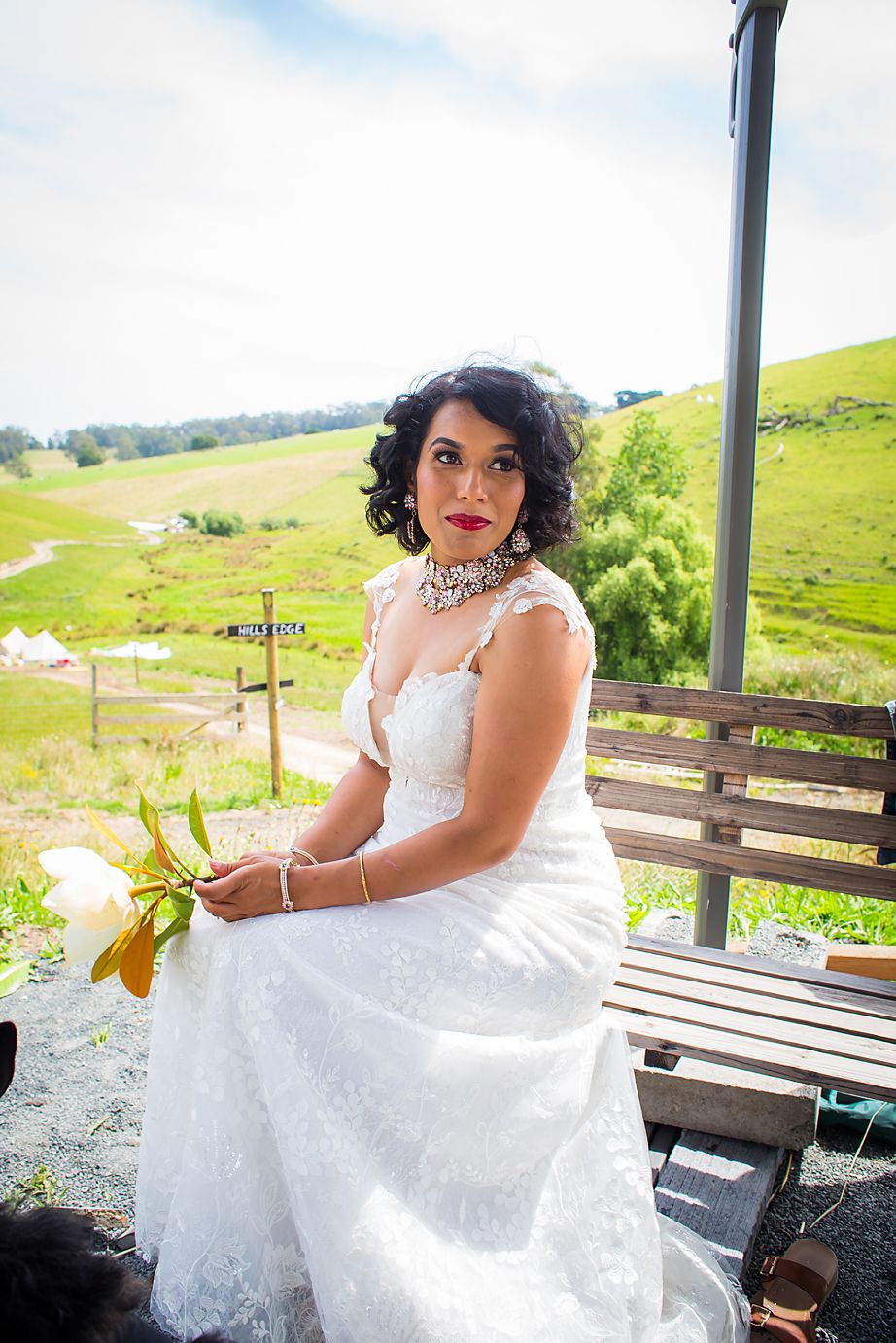 A person in a lace wedding dress poses sitting outdoors on a rustic wooden deck overlooking rolling green hills.