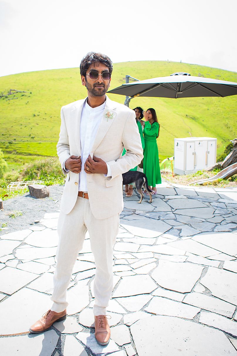 A groom in a white suit and sunglasses stands on a stone patio with green rolling hills in the background.