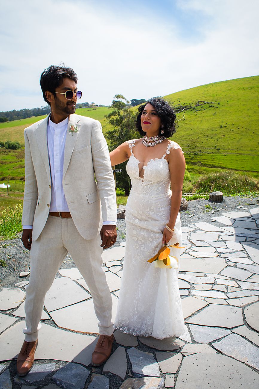 Wedding couple in white and beige attire share candid moments on a stone patio with green hills in background.